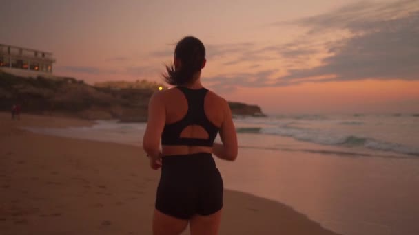 劇的な太陽の背景スローモーションでビーチで実行されている髪を振って運動女性 日没後のバックビューで海辺で屋外の物理的な活動を楽しんでいるスポーツ女性 後を追え — ストック動画