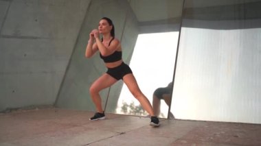 Çekici, orta yaşlı bir kadın, yan hamleler yapıyor, dışarıda egzersiz yapıyor. Dişi sporcu kasları ısıtıyor ve ayna duvarının yanında dikilirken esniyor. Kendine güven sportif kadın eğitimi