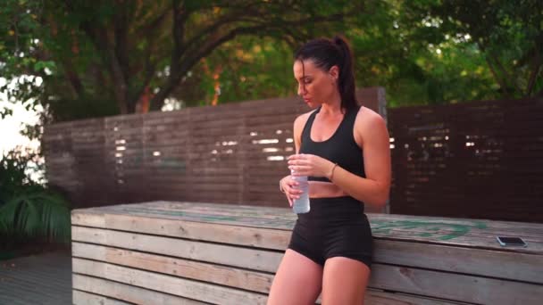 ジムの後のペットボトルから水を飲む中世の女性 屋外ワークアウト 都市スポーツ ブルネットの白人の女の子は暖かい夏の日市公園でトレーニングをジョギングした後 明確なミネラルウォーターを飲む — ストック動画