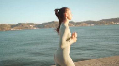 Confident fit female jogging by embankment area coastal city in beautiful sunny day do fitness training for cardio health. Tracking shot. Slow motion. Handheld. Athletic woman running, morning run