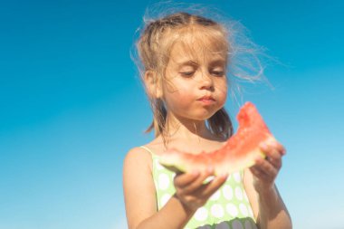 Funny little girl eat watermelon sunny summer day at ocean beach. Cute caucasian female child enjoy summer fruit bite slice of watermelon. Happy childhood. Close up portrait