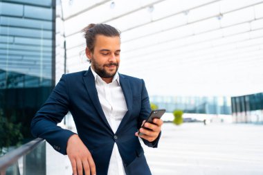 Açık havada duran bir iş adamı akıllı telefon görünümlü cihaz ekranı kullanır ve gülümser. Yakışıklı, İspanyol, iş adamı, orta yaşlı, takım elbiseli, akıllı telefonu elinde tutan. Modern iş adamı
