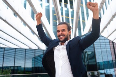 Excited businessman in suit celebrating victory arms raised up. Hispanic male business person winner pose. Best deal ever. Business man expressing positivity standing outdoors office building clipart