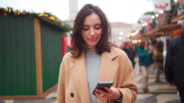 クリスマスマーケットでスマートフォンを使う女性 冬休みを楽しむ白人女性 ソーシャルネットワークの概念 アプリを使用して 背景にはクリスマスライトが点灯しています 肖像画 — ストック動画
