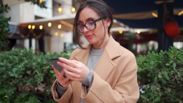 女性は屋外でスマートフォンを使う 秋の日 服を着たクリームコートの外に立つスマートフォンのデバイス画面を使用してメガネの肖像画の原因アジアの女性 人々はテクノロジーを使用しています — ストック動画