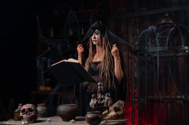 Karanlık zindan odasında duran siyah başlıklı cadı büyü yapmak için sihirli kitap kullanıyor. Kadın büyücü okültist medyum çekimi