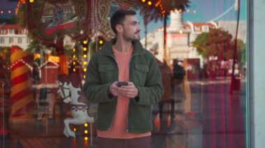 Portreci adam elinde akıllı telefonuyla dükkan penceresinin yanında duruyor ve kameraya gülümsüyor. Avrupa şehrinde çekici bir İspanyol adam. Şehir sokağı vitrinlere yansıdı