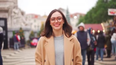Gözlüklü kadın şehir meydanında yürürken kameraya bakıp gülümsüyor. Gözlüklü Avrupalı kadın sıcak bir kış günü şehirde yürüyor.. 