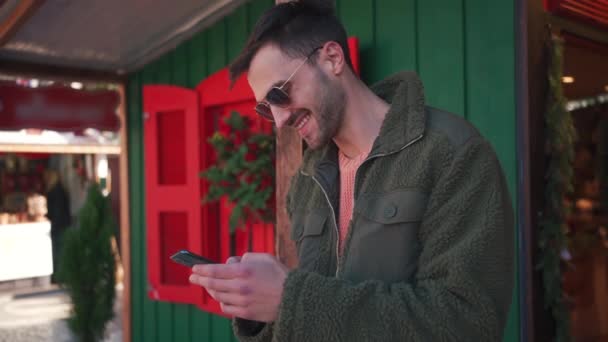 英俊而快乐的白人男子站在装饰过的拥挤的城市里 用智能手机发短信 并接受节日的问候 在圣诞节期间 年轻的成年男子在户外使用智能手机 — 图库视频影像