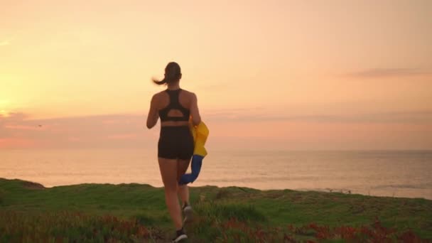 Αθλητική Γυναίκα Τρέχει Στη Θάλασσα Ηλιοβασίλεμα Κρατώντας Σημαία Της Ουκρανίας — Αρχείο Βίντεο