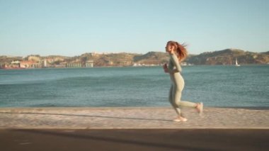 Confident fit female jogging by embankment area coastal city in beautiful sunny day do fitness training for cardio health. Tracking shot. Slow motion. Handheld. Athletic woman running, morning run