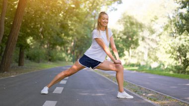 Kadın koşucu, yaz parkında egzersiz yapmadan önce bacak esnetiyor. Orta yaşlı atletik kadın, koşuya çıkmadan önce vücudu ısıtıyor. Beyaz şortuyla koşuyor.