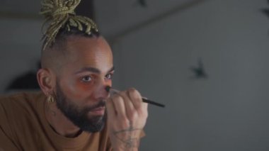 Black brazilian gay applaying make up eyeshadow looking mirror view from behind. Non binary androgynous guy. LGBT concept Close up face