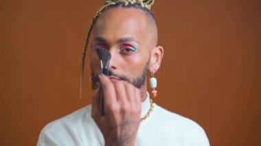 Homosexual man who applies powder to his face makeup brush. LGBT community with freedom for homosexuals. Black Brazilian gay applying make up looking camera standing isolated on brown background