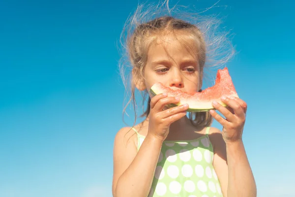 Funny little girl eat watermelon sunny summer day at ocean beach. Cute caucasian female child enjoy summer fruit bite slice of watermelon. Happy childhood. Close up portrait