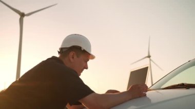 Technician engineer working at wind turbine using laptop computer worker or operator checks wind turbine farm does maintenance to power generator station lean on car wind turbine and sunset background