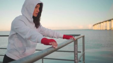 Athlete female doing push up exercises outdoor seascape background leaning on fence dressed hoodie sportswear. European athletic female practicing workout day. Slow motion close up. Fitness motivation