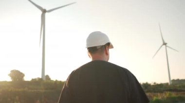 Engineer man in white hard hat walking on field with huge windmills during summer sunset. Professional technician. Portrait of an engineer wind turbines. Windmill engineer watching wind turbines 