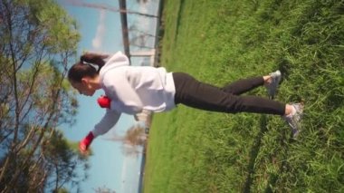 Elleri kırmızı boks kasetlerine sarılı, parkta boks yapan bir kadın. Orta yaşlı bayan sporcu kapüşonlu giyinmiş olarak dışarıda antrenman yapıyor. Dikey video. Atletik kadın boksör eğitimi.