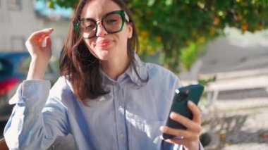 Woman in glasses using smartphone bench correct hairstyle outdoor looking on device screen and smile. Happy female wears casual clothes sit reads message or social media. Close up portrait