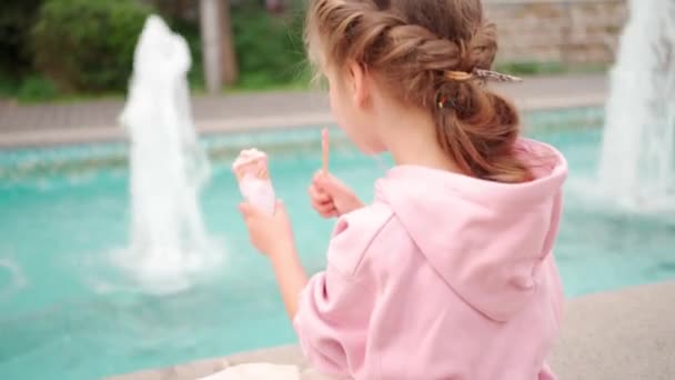 ピンクのかわいい女の子はおいしいアイスクリームを楽しんでいます 噴水の近くに座っているシティパークでアイスクリームを持つ子供 夏と子供時代を楽しむ — ストック動画