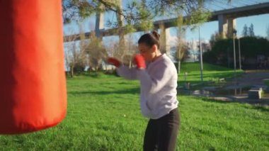 Woman shadow boxing with her hands wrapped in red boxing tapes with ocean bridge and blue sky background. Middle age female athlete fighter training outside dressed hoodie.