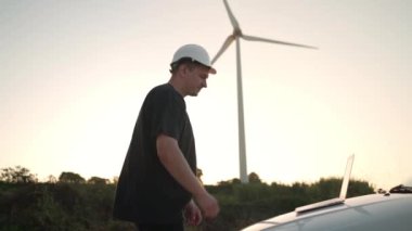 Technician engineer working at wind turbine using laptop computer worker or operator checks wind turbine farm does maintenance to power generator station lean on car wind turbine and sunset background