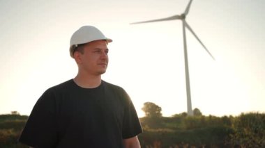 Engineer man in white hard hat walking on field with huge windmills during summer sunset. Professional technician. Portrait of an engineer wind turbines. Windmill engineer watching wind turbines 