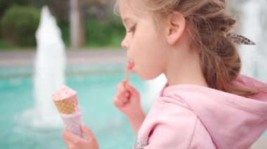 Little Cute Up Girl with Make in Pink Hoodie Enjoying Delicious Ice Cream. Kid with Ice Cream in the City Park Sitting near Fountain. Enjoy Summer and Childhood