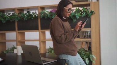 Businesswoman using smartphone in office. Professional female employee in glasses holding mobile phone in hand scrolling on device screen. Business woman dressed casual clothes in office meeting room