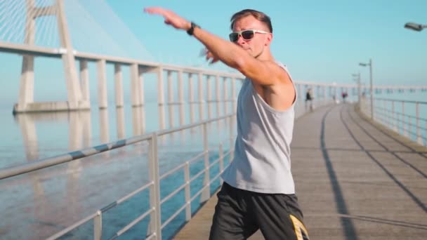 运动员在慢跑前热身 男子伸展胳膊和胸部 然后在室外跑步训练站在桥上夏季蓝天背景慢动作中等射门 — 图库视频影像