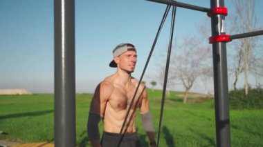 Erkek sporcu triseps kaslarını elastik bant ile çalıştırıyor. Gömleksiz atletik adam elastik direnç bandıyla triseps egzersizi yapıyor. Lastik bant sabit yatay çubuk. Spor, spor, spor.