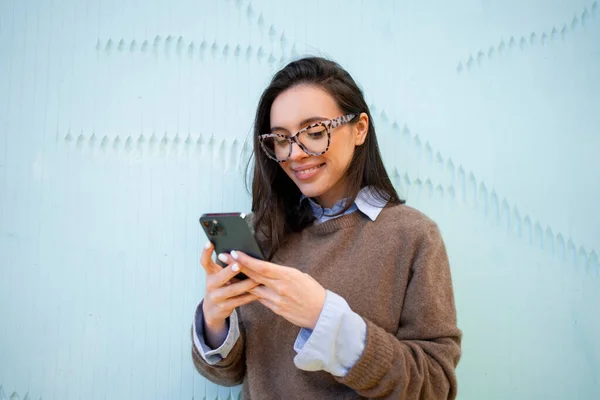 Satisfied hipster girl wear glasses, types text message on mobile phone, enjoys online communication, types feedback, wears sweater, isolated on blue color background. Technology concept