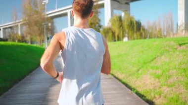 Erkek koşucu, güneşli bir parkta koşuyor. Şehir parkında kardiyo antrenmanı yapan bir atlet. Sabah sağlıklı yaşam tarzında gerçek insanları eğitmek. Çekimi takip et.