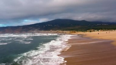 Bulutlu bir yaz gününde, Cascais, Portekiz 'deki Guincho Plajı' nda sörfçülerin ve sahile gidenlerin hızlı hava araçları görüntüsü. Dramatik gökyüzü, dağ plajı ve okyanus