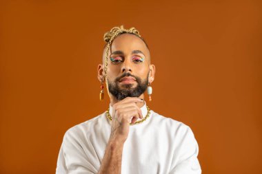 Kendine güvenen sakallı, parlak makyajlı eşcinsel adam turuncu arka planda izole edilmiş, beyaz giyinmiş. Gurur ve bireysellik hissi yayıyor. Kişisel tarzın çeşitlilik gücü