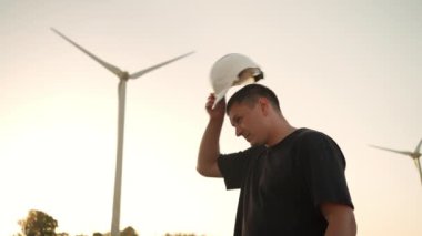 Engineer man in white hard hat walking on field with huge windmills during summer sunset. Professional technician. Portrait of an engineer wind turbines. Windmill engineer watching wind turbines 