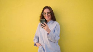 Sarı arka planda duran akıllı telefon kullanıcısı. Gözlüklü, elinde cep telefonu tutan, ekranda gülümseyen pozitif kadın.. 
