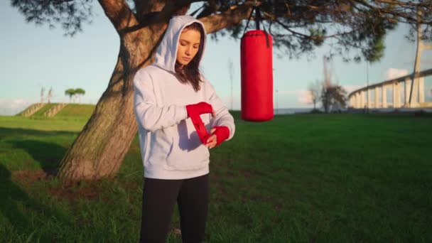 パンチングバッグの近くの公園に立つ女性ボクサー ボクシングで腕の強力で自信のある女性の周りのカメラの包装ボクシングテープを見て 女性戦闘機 強い女性と自己防衛の概念 — ストック動画