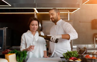 Yakışıklı genç Afrikalı şef, beyaz kız arkadaşıyla mutfakta kırmızı şarap malzemesi kullanarak yemek pişiriyor. Bir aşçı bir kıza nasıl yemek yapılacağını öğretir. Erkek ve kadın profesyonel mutfakta yemek pişiriyor..