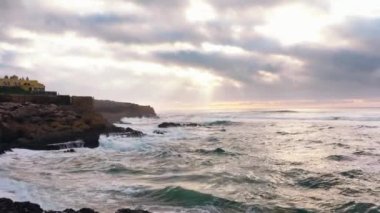 Gün batımında okyanusun üzerinden uç. Bulutlu bir yaz gününde, Portekiz Praia da Guincho 'daki Atlantik Okyanusu sahilinde bir otel Praia da Guincho hava manzaralı. Dramatik gökyüzü, dağ plajı ve okyanus. Sinematik görünüm. 