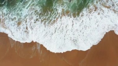 Kumlu sahil şeridinde dalgalanan turistik deniz dalgalarının güzel, pürüzsüz görüntülerini gösteren drone görüntüsü. Altın plajın havadaki görüntüsü derin mavi okyanus suyu ve köpüklü dalgalarla buluşuyor.