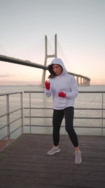 Dedicated female boxer dreesd hoodie warming up arm muscule. Athletic female prepares for boxing workout outdor with city seascape. Female fitness motivation. Fighter warm up body before training 