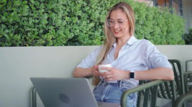 İş kadını internet üzerinden toplantı için video görüşmesi yapıyor ve tavalarını gülen yüzle sunuyor ve gülüyor. Kahve fincanını tut. Serbest çalışan bir kadın, internetteki video görüşmesi için oturmakta olan kafe terasında laptop kullanıyor.