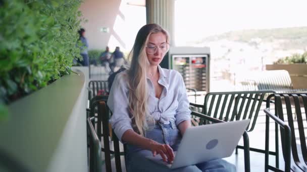 带笔记本电脑的自由职业者坐在室外咖啡厅的平台上 在室外使用笔记本电脑戴眼镜的金发女性 中镜头高角镜 使用笔记本电脑从事远程工作的妇女 — 图库视频影像