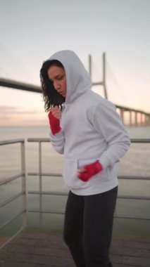 Female boxer dreesd hoodie warming up arm muscule. Athletic female prepares for boxing workout outdor with city seascape.Fighter warm up body before training Female fitness motivation. Outdoor gym