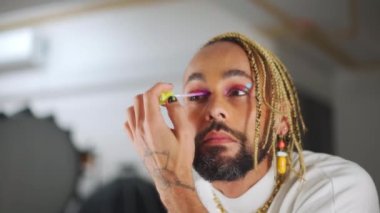 Male Caucasian extravagant transsexual man applying mascara on eyelashes. Close-up portrait of confident male queer doing makeup standing near mirror. Cosmetics and gender identity