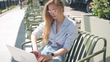 Açık kafe terasında dizüstü bilgisayarı olan serbest çalışan biri. Dışarıda dizüstü bilgisayar kullanan gözlüklü sarışın bir kadın. Orta açı, yüksek açı görüntüsü. Uzak görev için dizüstü bilgisayar kullanan kadın.