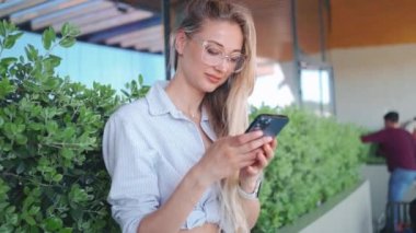 Neşeli, beyaz bir kadın. Gündüz vakti telefonda mesaj yazıyor. Sarı saçlı, gülümseyen gözlüklü ve tişörtlü. Sosyal medya bağımlılığı kavramı