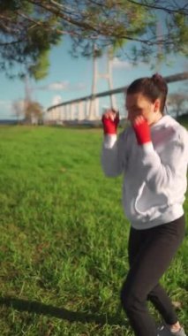 Elleri kırmızı boks kasetlerine sarılı, parkta boks yapan bir kadın. Orta yaşlı bayan sporcu kapüşonlu giyinmiş olarak dışarıda antrenman yapıyor. Dikey video. Atletik kadın boksör eğitimi.